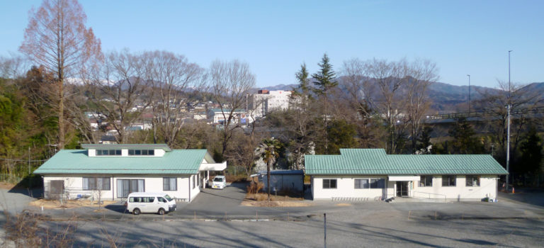 緑色の屋根を持つ平屋建ての介護施設の外観。駐車場と周辺の山々が見える。