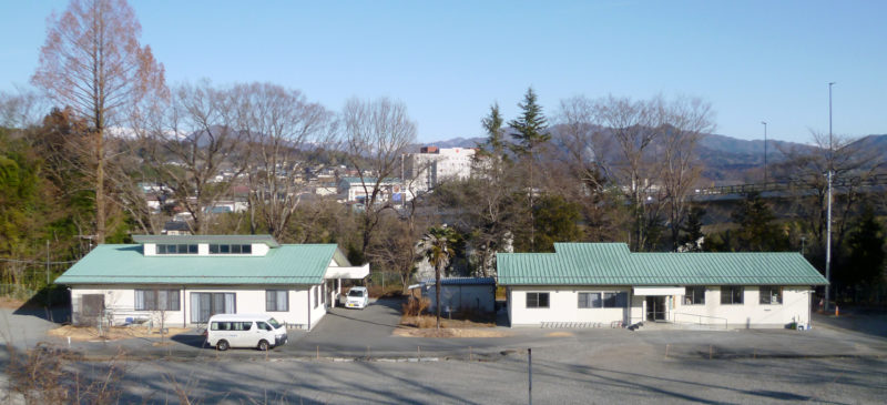 緑色の屋根の平屋建て施設。駐車場と周辺の山々が見える。